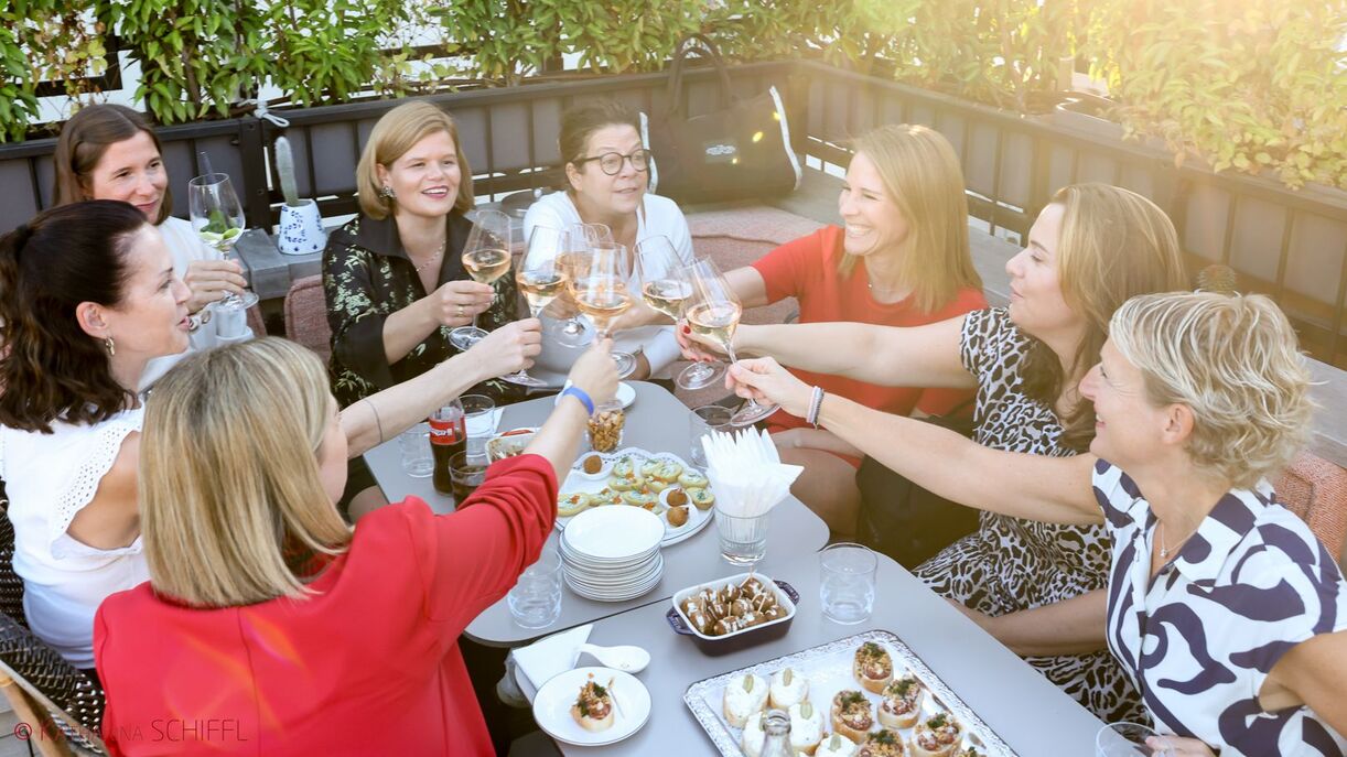 Acht Damen bei einem länglichen Tisch sitzend, die mit Gläsern anstoßen.