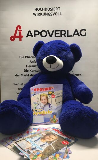 Brunner Teddyklinik APOLINO Brunner Teddyklinik APOLINO - Ein großer, blauer Teddybär sitzt mit APOLINO-Magazinen vor einem APOVERLAG-Roll Up.