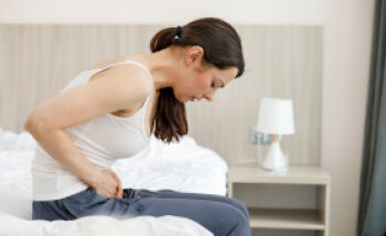 Webinar: FEMANNOSE®N bei Blasenentzündungen - Eine junge Frau sitzt aufgrund der Schmerzen einer Blasenentzündung mit nach vorne gekrümmtem Oberkörper auf dem Bett.